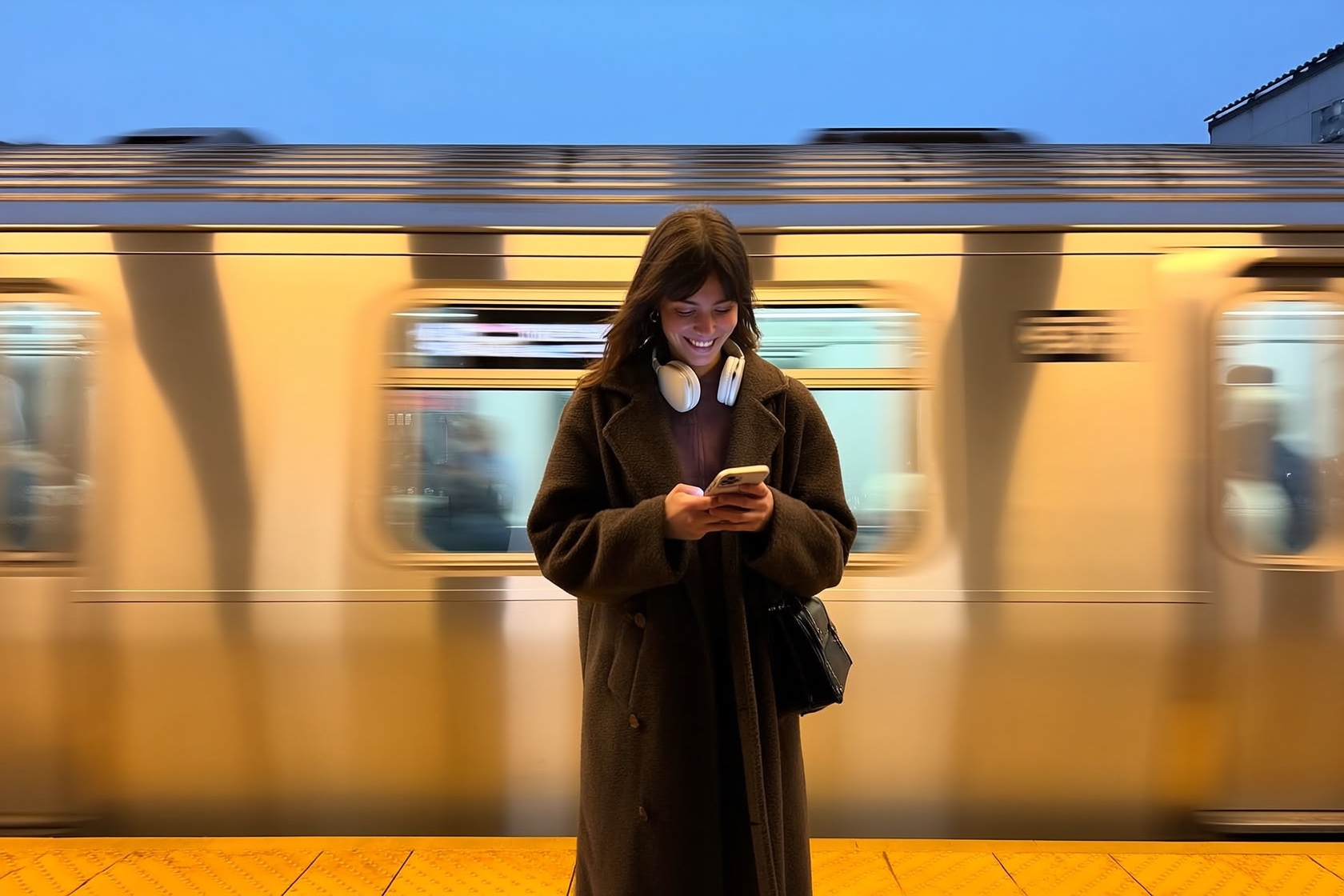 A commuter smiling at her phone on a subway platform as a train passes behind her.