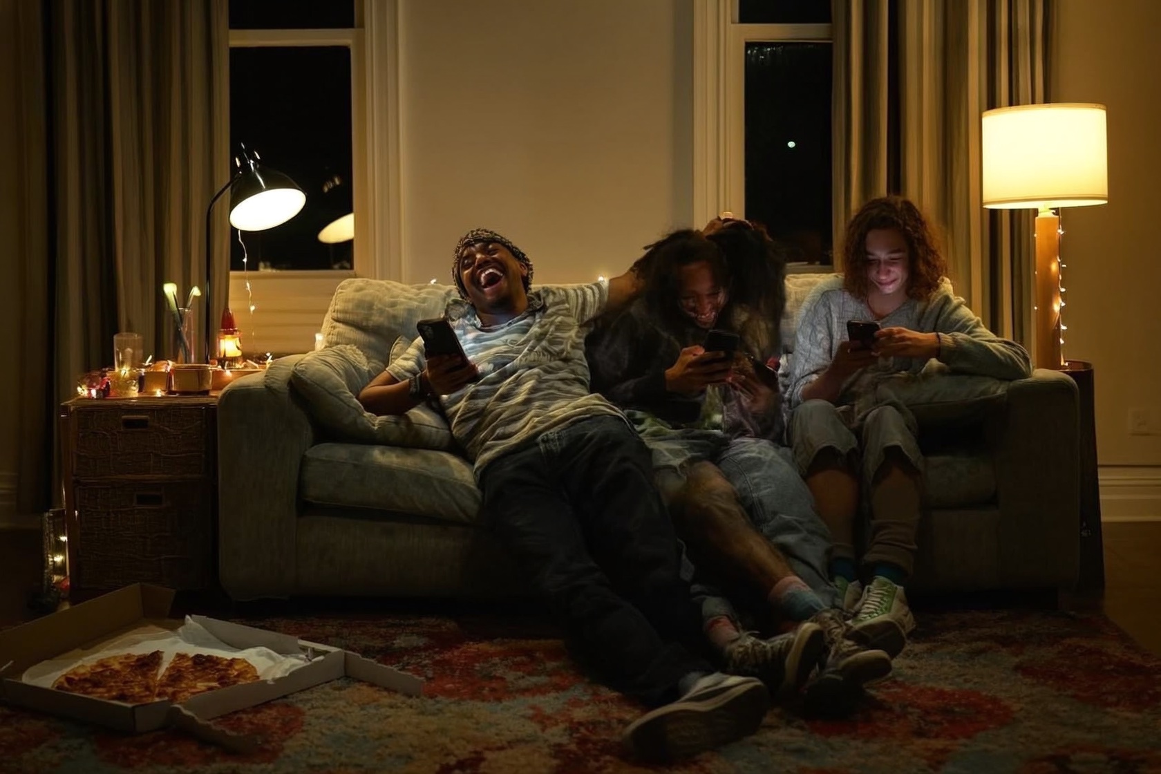 Three friends on a sofa at night, phones in hand, one laughing with his head thrown back.